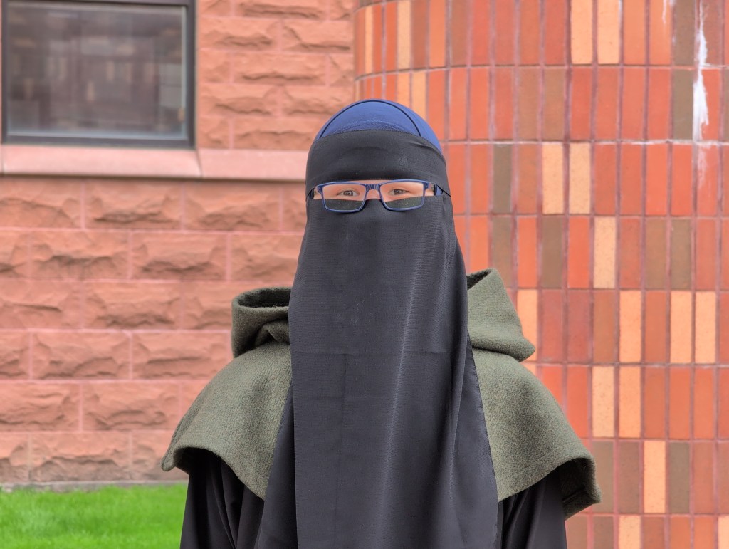 Euphoria Rising is a Muslim trans woman wearing a blue hijab, a black niqab (face veil), an army green druid hood, and a black abaya. This photo was taken against a reddish brown brick wall.